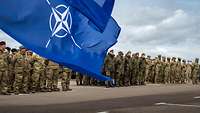 NATO- und EU-Flagge wehen im Wind, im Hintergrund angetretene Soldaten unterschiedlicher Nationen