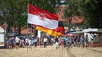 Besucher bei einer öffentlichen Veranstaltung im Vordergrund die Fahnen des Landes Brandenburg und Deutschlands.