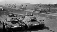 Schwarz-Weiß-Aufnahme von Panzern und Hubschraubern im Gelände