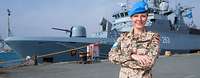 Eine deutsche Soldatin mit blauem UN-Barett steht vor einer Fregatte im Hafen