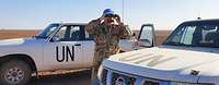 Ein deutscher Soldat mit blauem Basecap steht zwischen zwei weißen UN-Fahrzeugen und schaut durch ein Fernglas
