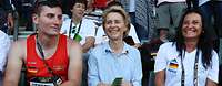 Von der Leyen sitzt auf der Tribüne zwischen einem Sportler und einer Frau