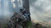 Ein Soldat kniet mit Waffe im Anschlag hinter einem Baum im Wald