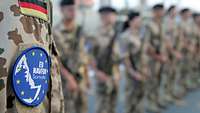 Nahaufnahme von einem Patch „EU NAVFOR Somalia“ auf einer Soldatenuniform. Im Hintergrund angetretene Soldaten an Deck.