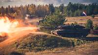 Ein Kampfpanzer Leopard mit niederländischer Flagge schießt scharf im Gelände. Aus seinem Rohr strömt ein Feuerball.