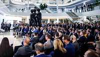 Viele Personen sitzen in einem Foyer und hören einer Podiumsdiskussion zu.