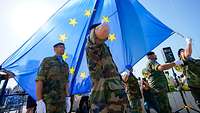 Mehrere Soldaten verschiedener Nationen hissen eine große EU-Flagge während einer Zeremonie.