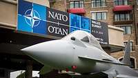 Ein Eurofighter steht vor einem Gebäude, im Hintergrund ein Plakat der NATO