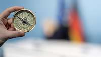 A hand holds a compass into the picture. In the background, you can see a blurred EU and German flag