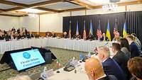 Christine Lambrecht sitzt mit hochrangingen Politikern in einem Konferenzsaal. Im Hintergrund sind Flaggen der USA und Ukraine.