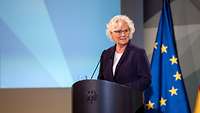 Ministerin Lambrecht steht am Rednerpult, dahinter die Flagge der EU.