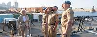 gerade tritt Ministerin Lambrecht an Bord eines Militärschiffes, begrüßt von drei Soldaten, im Hintergrund ein Containerhafen