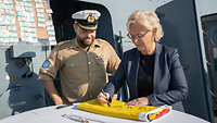 Verteidigungsministerin Lambrecht unterzeichnet eine zusammengefaltete Flagge, neben ihr steht der Korvettenkapitän.