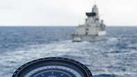 Ein Teil eines Kompasses ist zu sehen, dahinter unscharf ein Schiff der Marine.