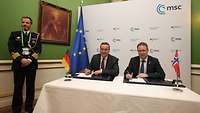 Boris Pistorius und Bjørn Arild Gram sitzen nebeneinander an einem Tisch und unterzeichnen eine Vereinbarung.