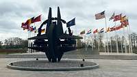 Die NATO Rose steht als Skulpture vor dem Hauptquartier der NATO im Hintergrund sind Flaggen der Mitgliedstaaten.