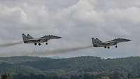Zwei Kampfflugzeuge fliegen hintereinander über Wald unter grau bewölktem Himmel