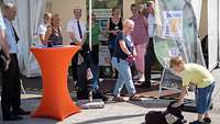 Mehrere Personen an einem Stand schauen auf ein Kind, das einen schwarzen Hund streichelt