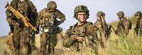 A soldier kneels between grass and holds a rifle in his hand. There are more soldiers in the background.