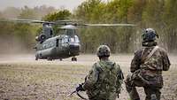 Zwei Soldaten hocken auf Feldboden und schauen in Richtung eines Transporthubschraubers vom Typ Chinook.