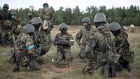 Deutsche und ukrainische Soldaten hocken auf offenem Gelände im Kreis um eine Sprengfalle und besprechen sich