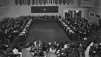 historisches Foto: in einem ein großer Saal, gekrönt vom Emblem der NATO, werden Politiker von Kamerateams beobachtet
