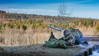 Drei Soldaten hocken auf Waldboden und schießen mit einer Panzerabwehrwaffe. Im Hintergrund ist ein Nadelwald erkennbar.