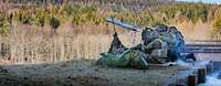 Drei Soldaten hocken auf Waldboden und schießen mit einer Panzerabwehrwaffe. Im Hintergrund ist ein Nadelwald erkennbar.