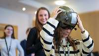 Eine junge Frau probiert einen Helm eines Marinefliegers aus. Hinter ihr stehen junge Frauen 