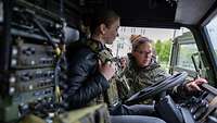 Eine Frau sitzt in einem Gefechtsfahrzeug mit Schutzweste, während eine Soldatin ihr alles erklärt