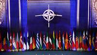Ein in blau beleuchteter, Raum ist zu erkennen. In der Mitte ist das Emblem der NATO platziert, davor stehen Flaggen.
