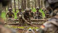 Vermummte Soldaten hocken auf Waldboden nah beieinander, während ein Soldat die restlichen Soldaten einweist. 