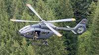 Über einen Tannenwald fliegt ein Leichter Kampfhubschrauber H145M. Die Aufnahme wurde aus der Luft gemacht.