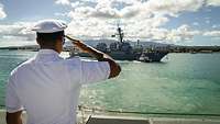 Von hinten sieht man einen Marine Soldaten, der von einem Pier aus militärisch grüßt. Vor ihm eine Fregatte.