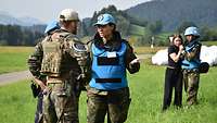 In a staged scene, several soldiers talk to each other, behind them a female soldier comforts a civilian.