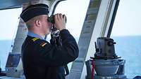 Ein Soldat steht auf einer Schiffsbrücke und schaut mit einem Fernglas aufs offene Gewässer.