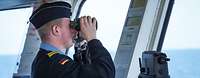 Ein Soldat steht auf einer Schiffsbrücke und schaut mit einem Fernglas aufs offene Gewässer.