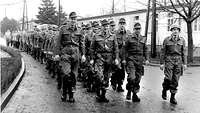 Schwarz-Weiß-Foto: Uniformierte Soldaten in Marschkolonne auf einem Kasernengelände